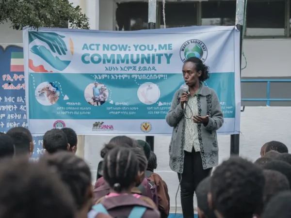 Dr Hanna stands in front of a crowd of children. Behind her is a big poster giving information on rabies prevention
